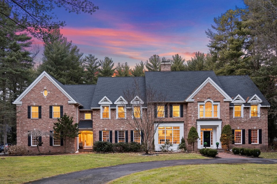 Large brick home with multiple gables and glowing windows among tall trees at dusk.