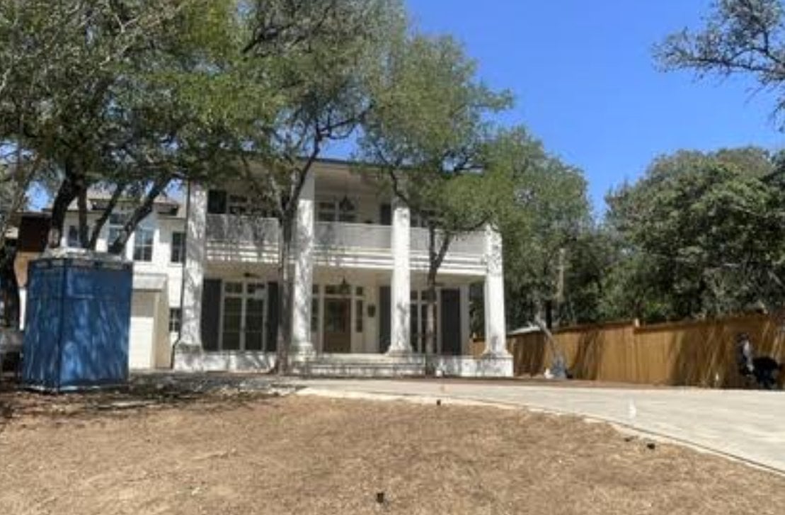 Two-story white house with a covered balcony, mature trees, and a blue portable toilet beside the driveway.