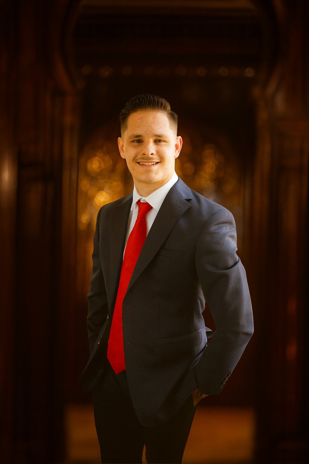 Professional portrait of a young male real estate agent in a navy suit and red tie, smiling and standing in front of a warm, elegant, golden-toned architectural background.