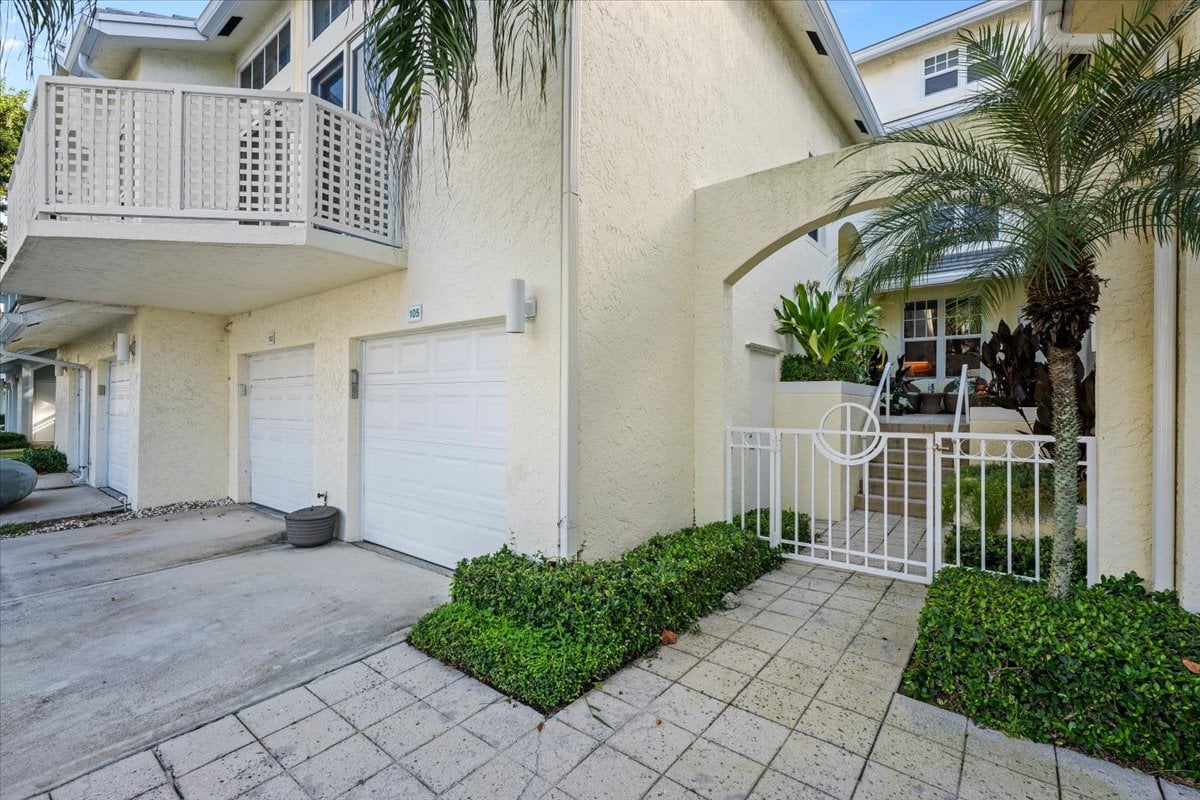 Cream stucco condominium entrance with a gated walkway, tropical palms, balcony, and garage doors.