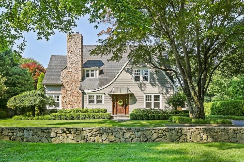 Gray-shingled house with a tall stone chimney, mature trees, and a stone wall along the front lawn.