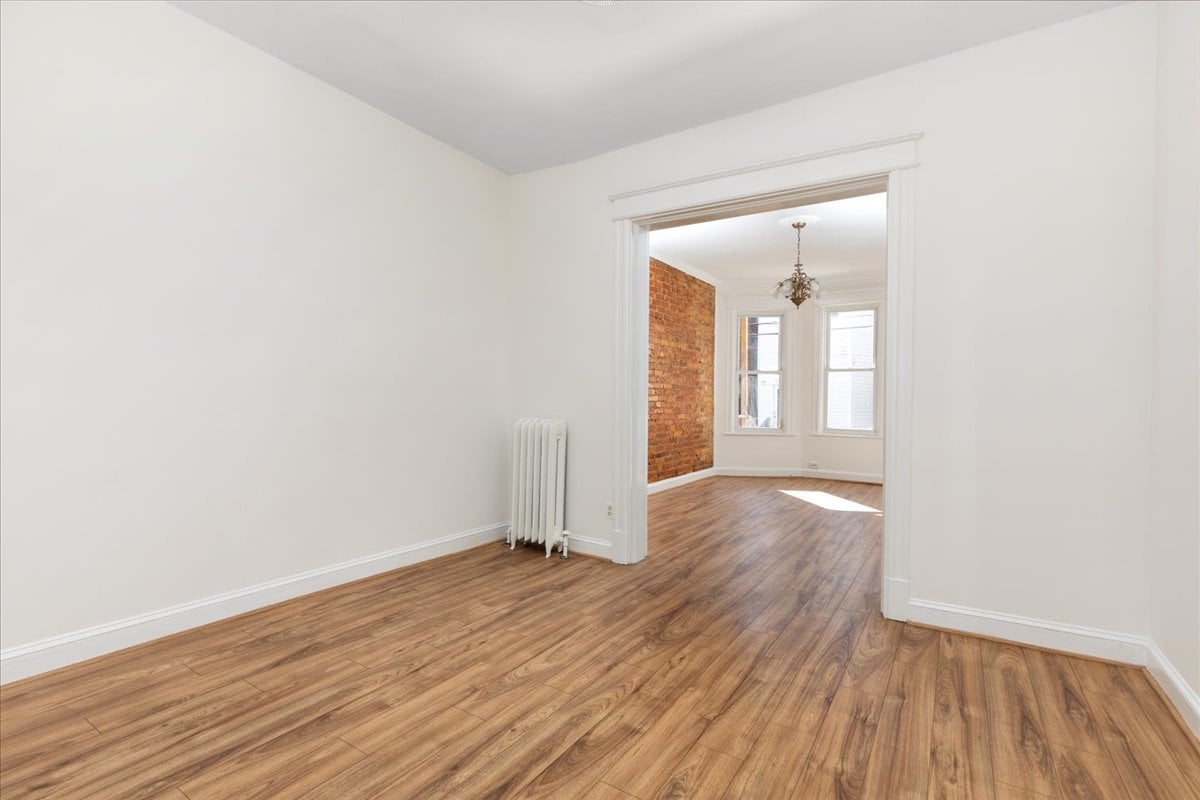 Empty room with hardwood floors and a radiator, opening to a sunlit room with exposed brick.