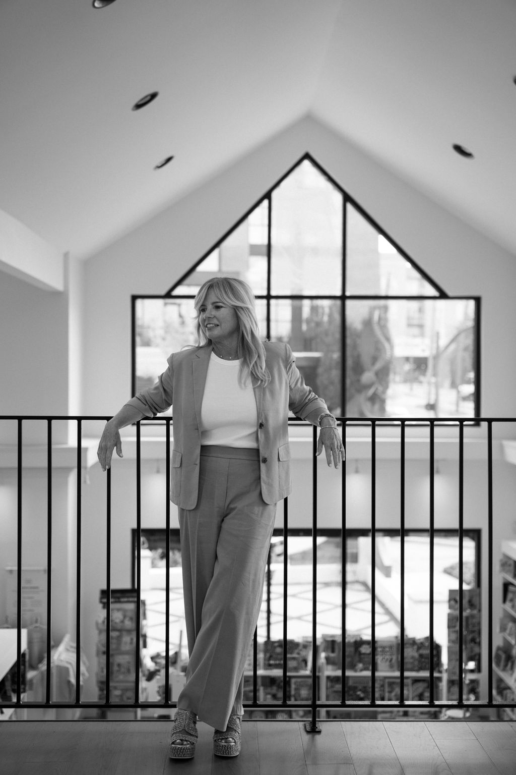 Blonde woman in a blazer leans on a railing before a large triangular window.