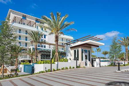 Modern condominium entrance with glass balconies, palm trees, and a paved entry plaza.