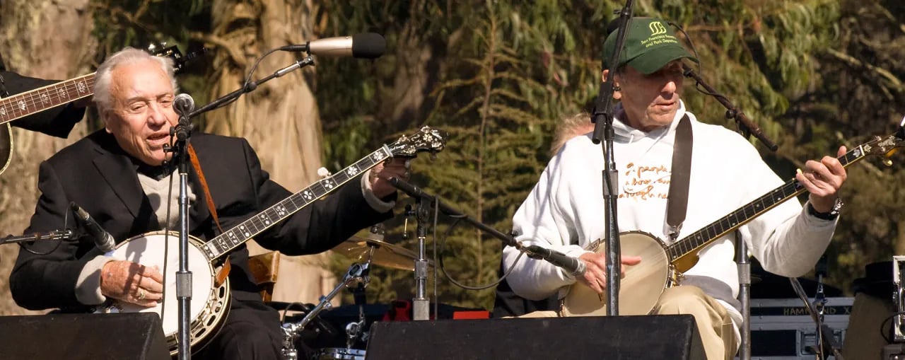 Two elderly musicians performing together on outdoor stage, both playing banjos, one in black suit and one in white t-shirt with green cap, with natural foliage backdrop