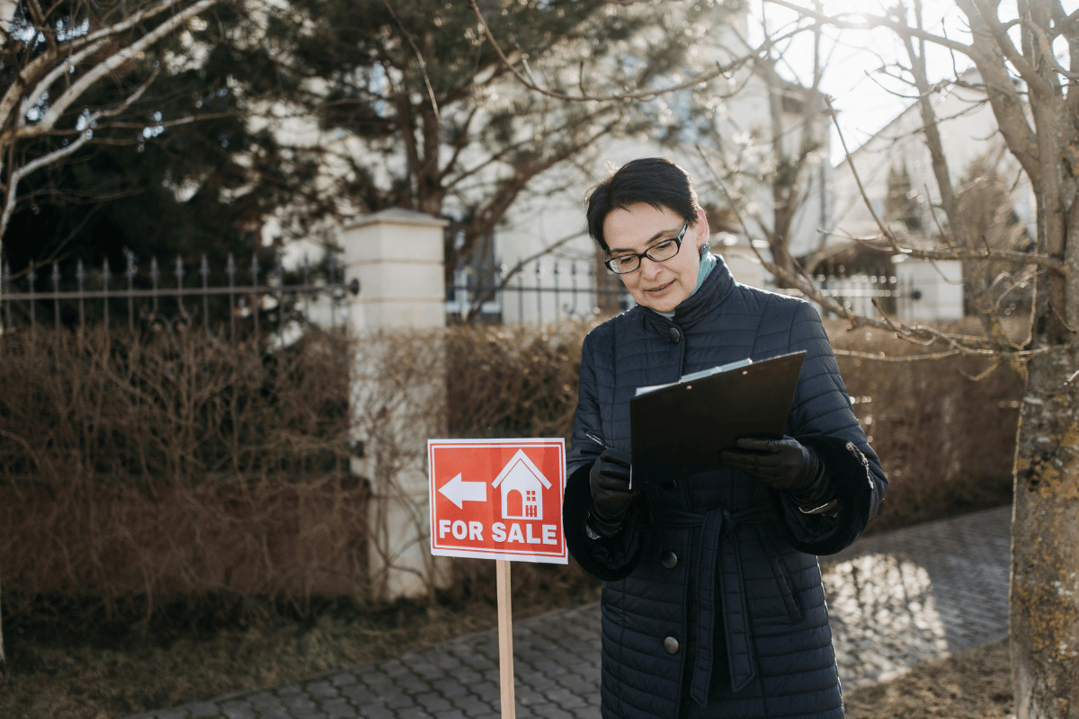 Real estate agent holding a clipboard near a “For Sale” sign in a winter setting.