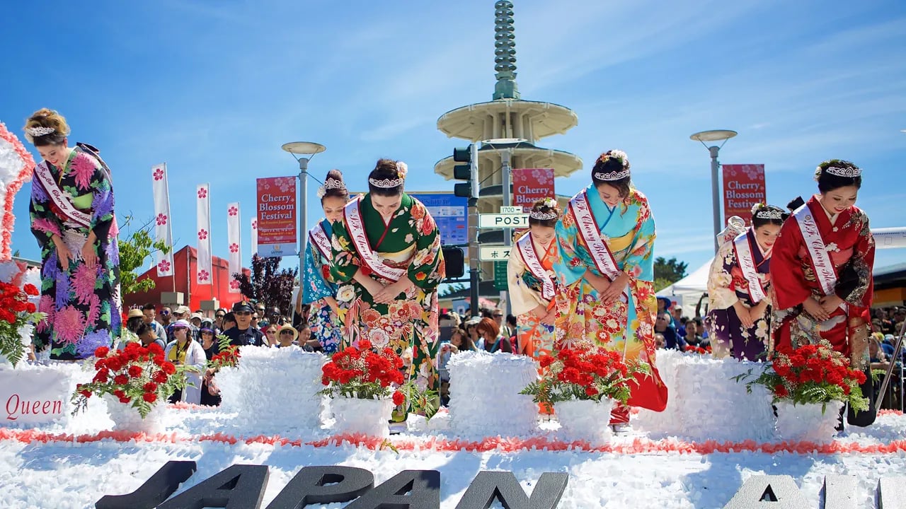 Women in traditional kimonos and tiaras bowing on a parade float at the 2016 Northern California Cherry Blossom Festival, with a pagoda in the background.