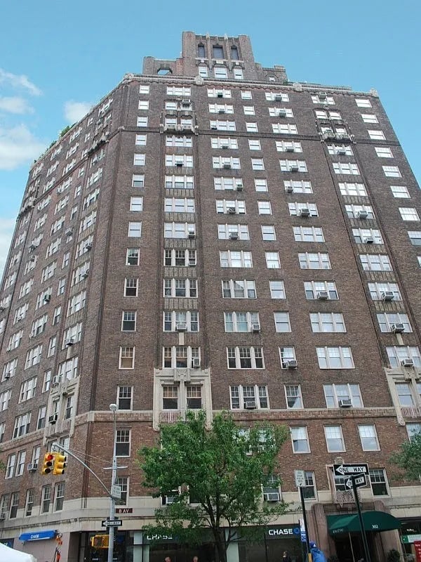 Tall brown brick apartment building with rows of windows and storefronts along the street.