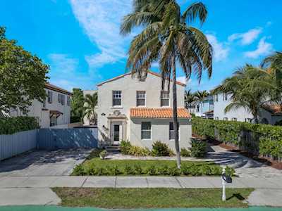 Two-story light-colored home with a terracotta awning, palm trees, and a paved driveway.