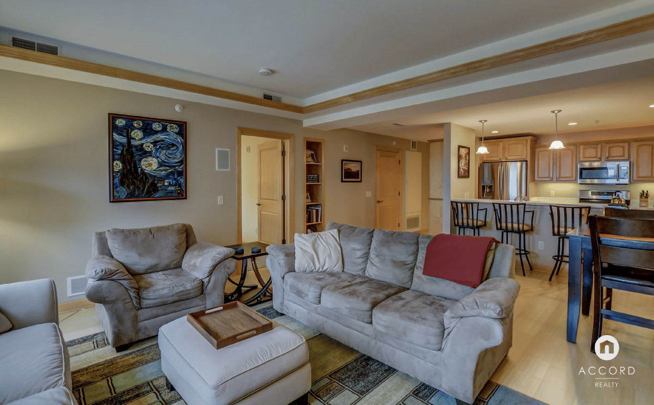Open-plan condo living room with gray sofas, patterned rug, and adjoining kitchen.