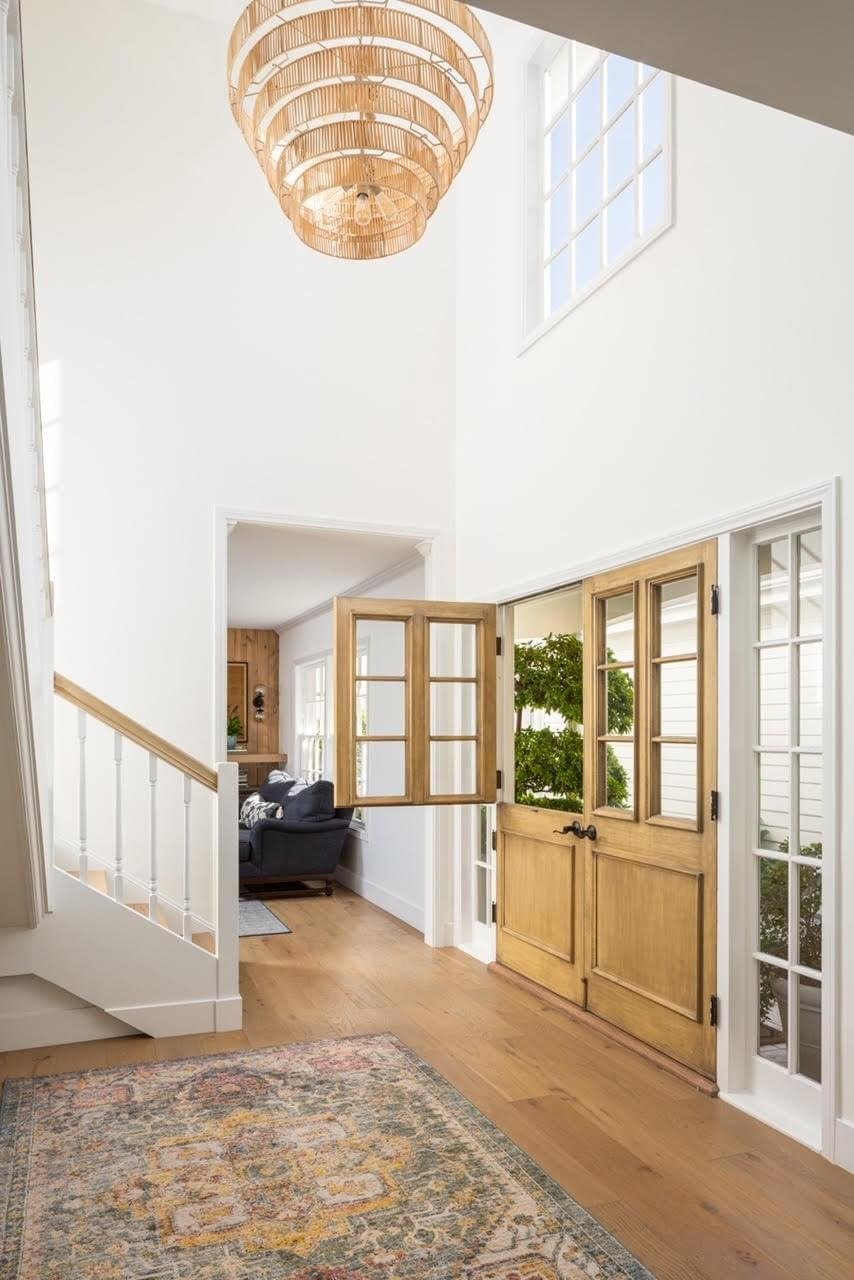  A home entryway with an open Dutch door, a large chandelier, and a colorful rug on the light wood floor.