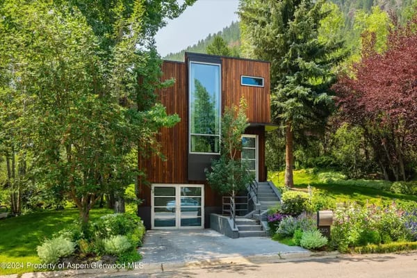 Modern wood-clad home with tall windows, landscaped gardens, and a forested mountain behind it.