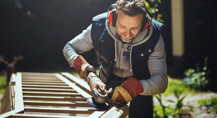 Man wearing protective gloves and earmuffs uses a power tool to sand a wooden structure outdoors. He appears focused and content. Bright, sunny day.