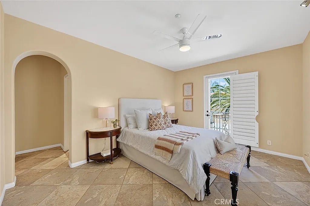 Staged bedroom with a white bed, tan tile floor, arched doorway, and shuttered balcony door.