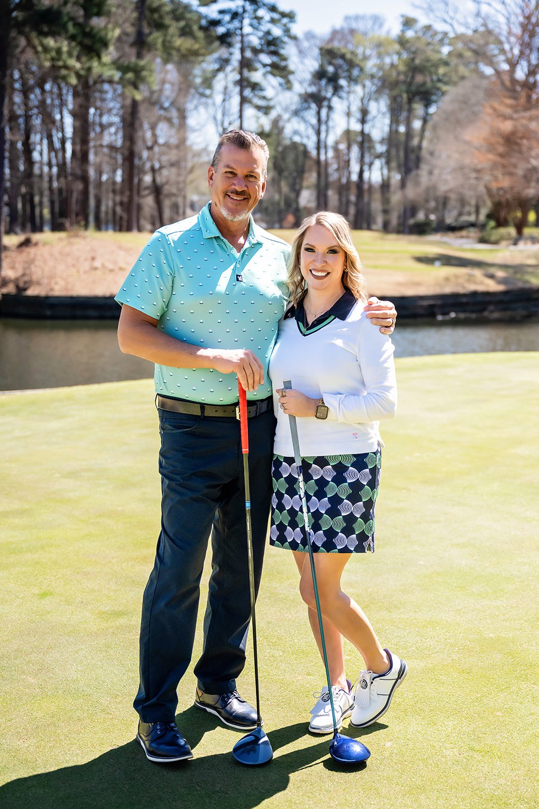 Heather Heishman and a man pose with golf clubs on a sunny golf course.