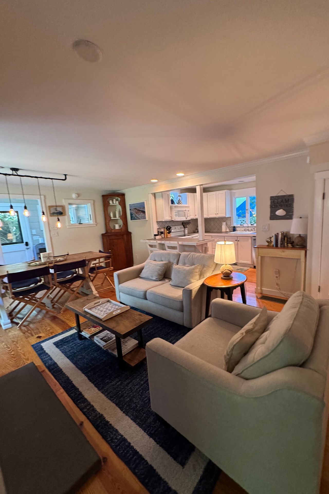 Open-plan living room with gray sofas, a dark blue rug, dining table, and white kitchen.