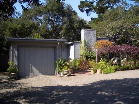 Home with a gray garage door, white chimney, and mature trees around the gravel driveway.