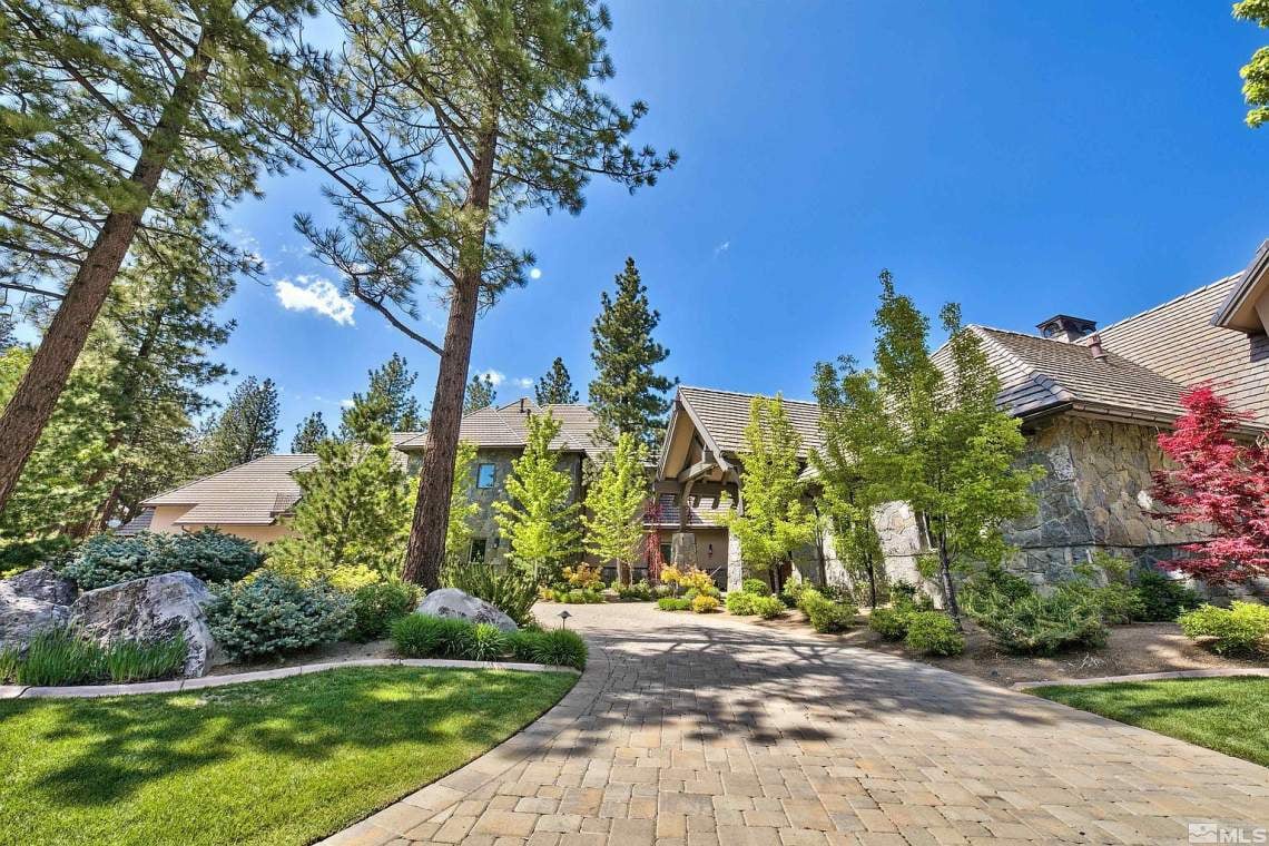 Large stone-and-wood home framed by pine trees, with a landscaped garden and broad paved driveway.