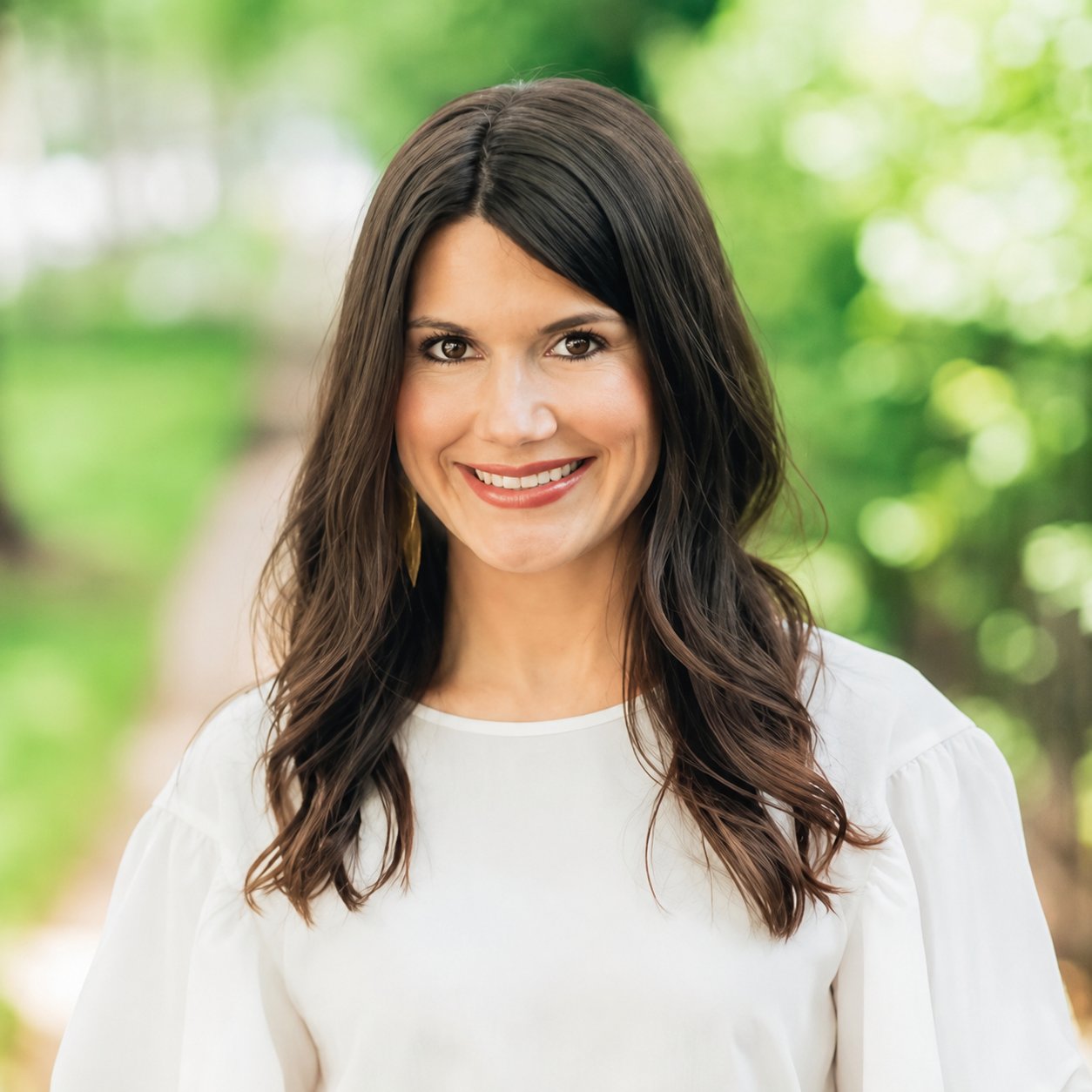 Smiling woman with long dark hair in a white blouse against a leafy background.