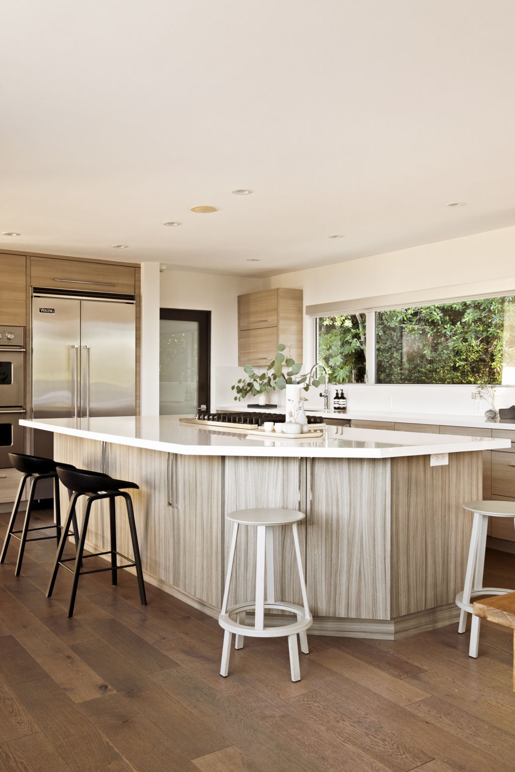 Modern kitchen with a large white island, pale wood cabinetry, and a window overlooking greenery.