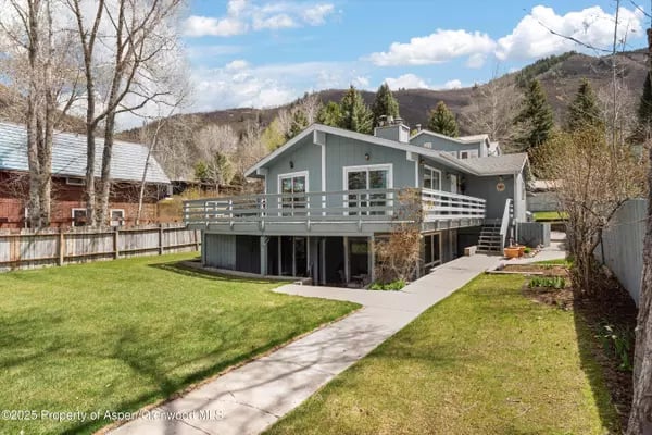 Gray two-story house with a broad deck, lawn, and wooded hills behind it.