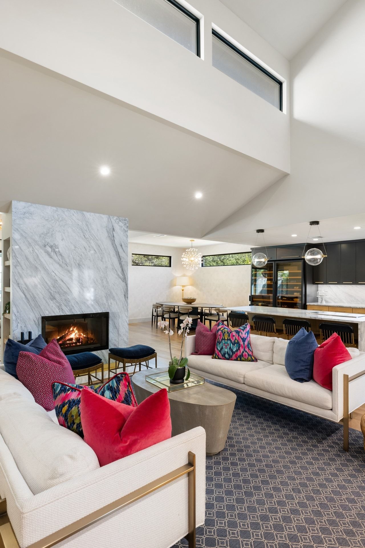 Open-plan living room with a veined marble fireplace, vaulted ceiling, and colorful furnishings.