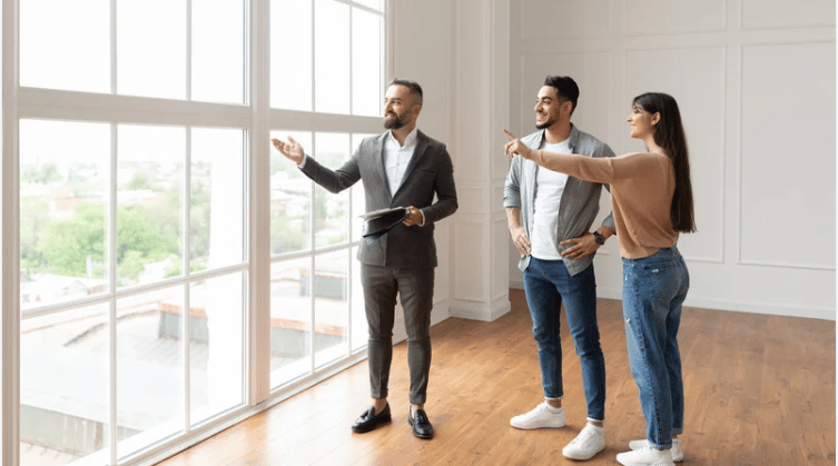 An agent shows a couple a bright, vacant room with tall windows and hardwood floors.