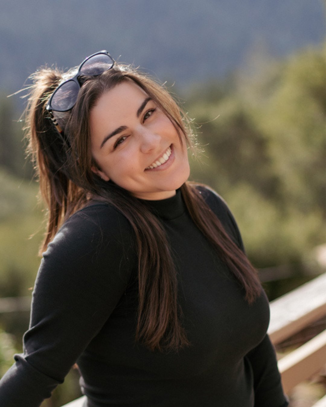 Smiling woman with long brown hair and sunglasses perched on her head, wearing a black top.