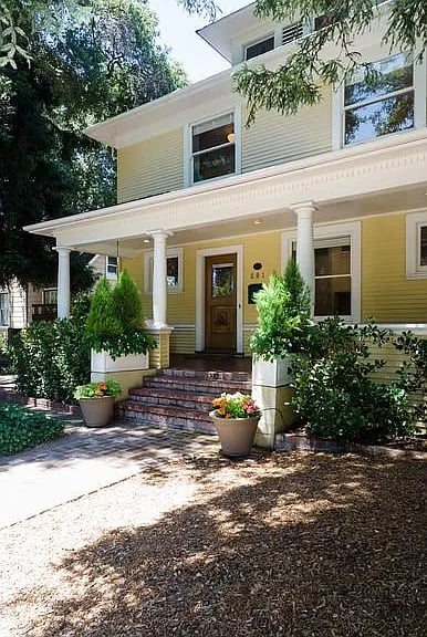 Yellow two-story house with a columned porch, brick steps, and potted flowers.