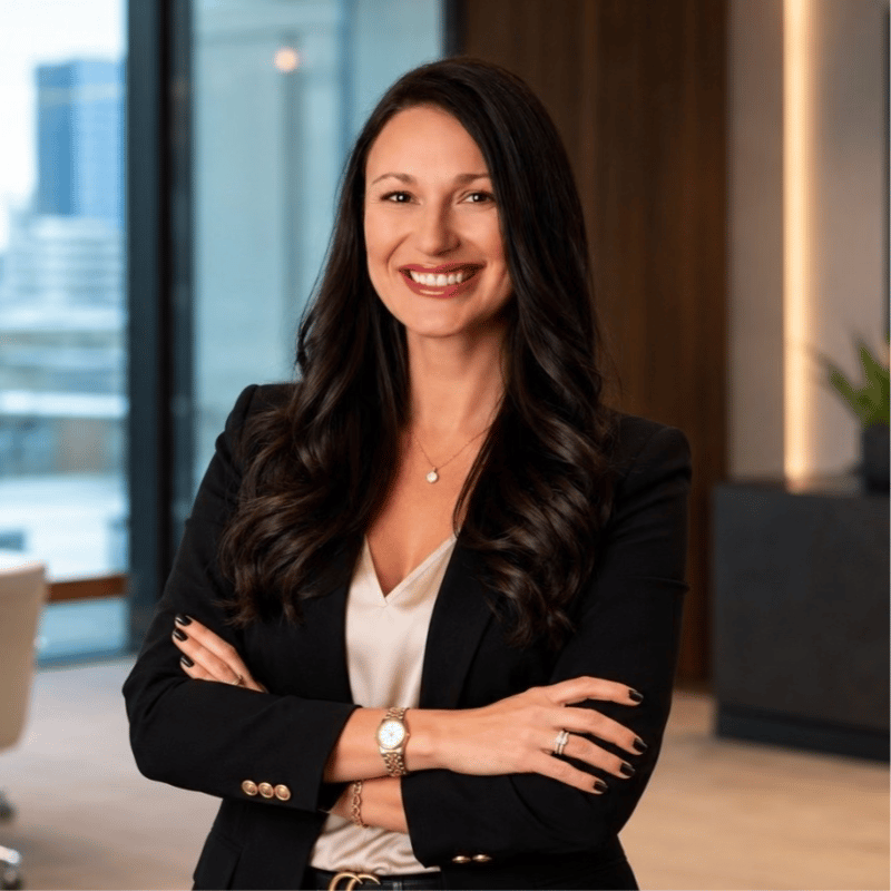Smiling woman with long dark hair in a black blazer, standing with arms crossed in an office.
