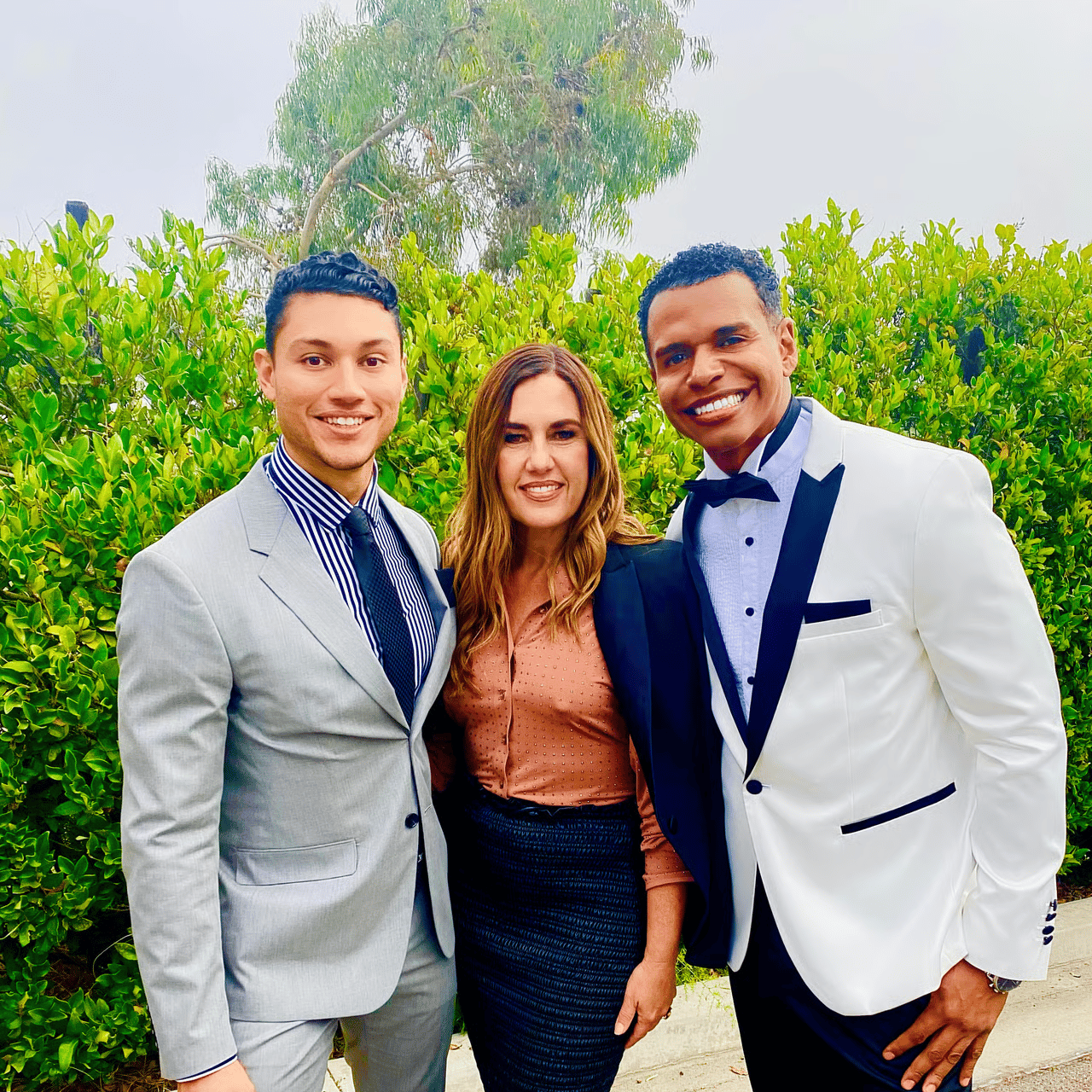 Three formally dressed adults pose together in front of a leafy hedge.