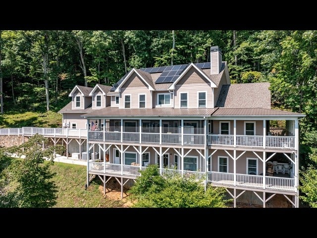 Two-story gray house with solar panels and wraparound decks among wooded surroundings.