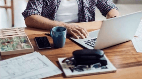 Person working at a wooden desk with a laptop, phone, mug, and papers.