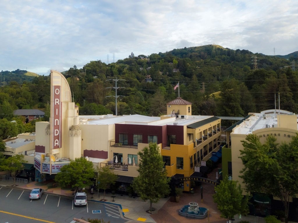 Elevated view of Orinda Theatre Square featuring the iconic Art Deco "ORINDA" marquee sign, multi-story Mediterranean-style retail shops, a central courtyard fountain, and lush green hills.