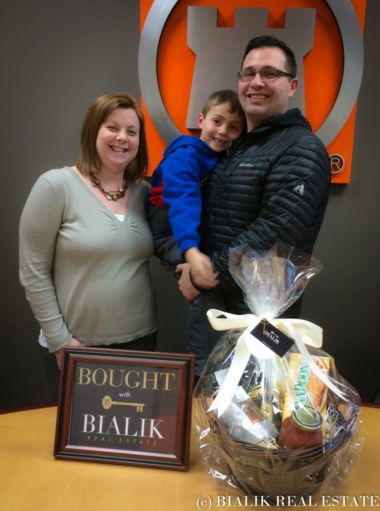 Kyle and Catherine pose with a child beside a Bialik Real Estate closing-day sign and gift basket.