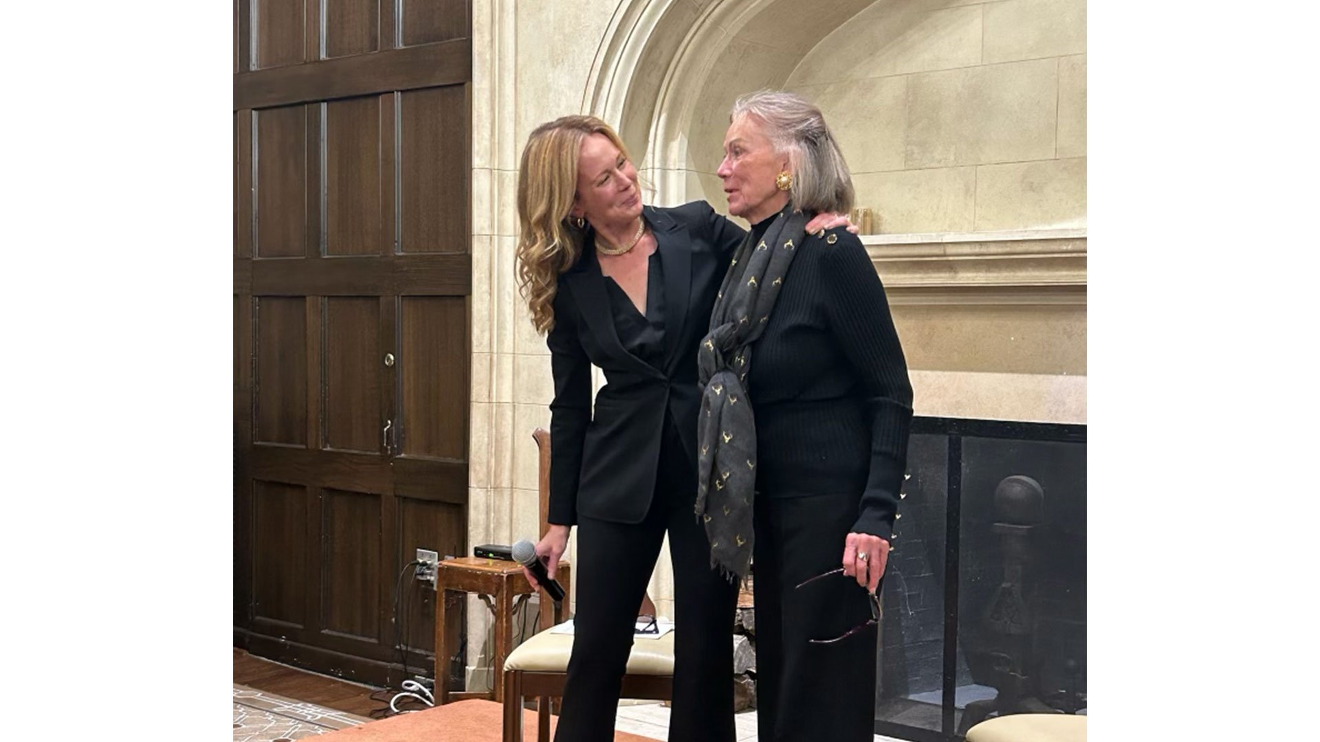 Two women are engaged in a warm conversation, one wearing a black suit and the other in a black outfit with a scarf, in a cozy indoor setting.