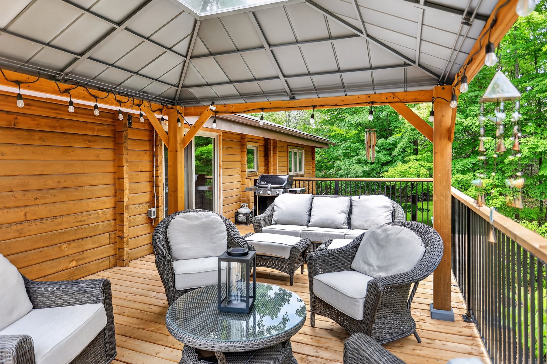 Covered cottage deck with wicker seating, string lights, and a barbecue beside leafy trees.