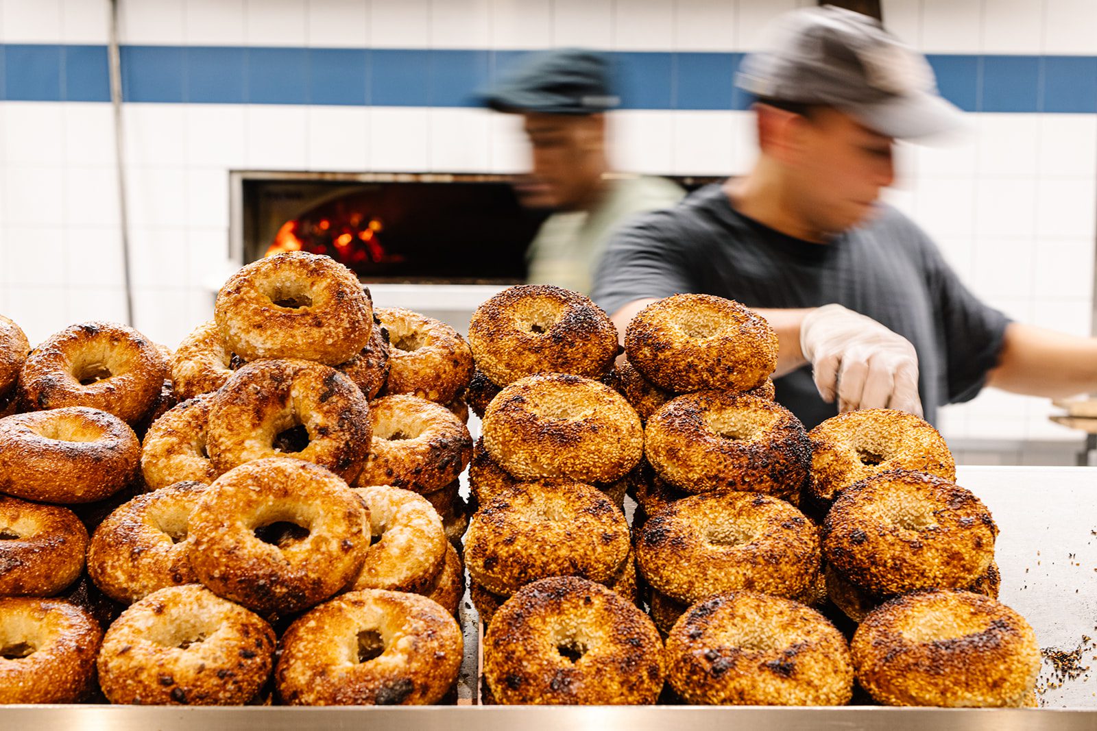 A display of freshly baked bagels stacked high, with bakers in motion in the background.