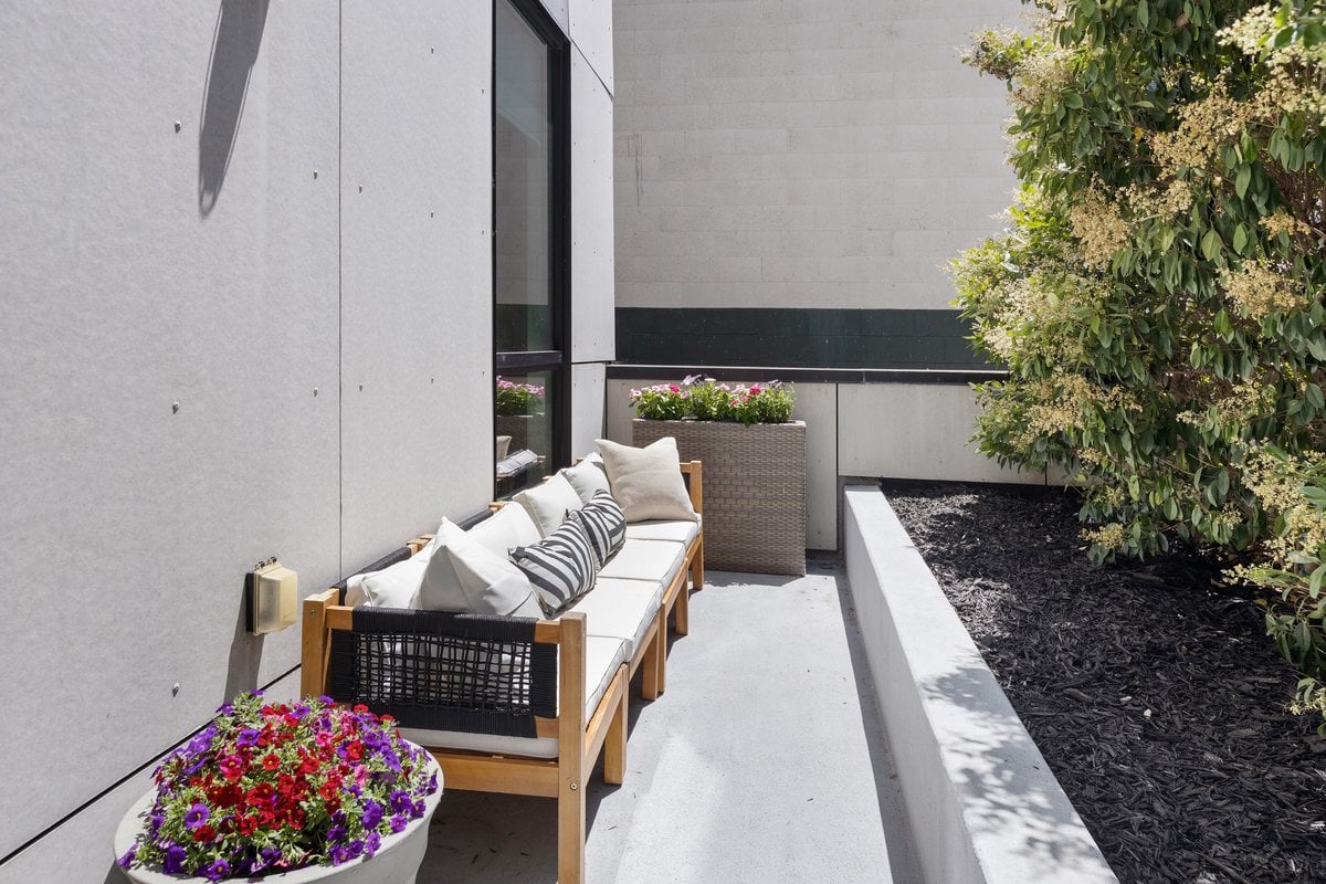 Patio with cushioned wood sofa, colorful flower planters, and raised garden bed beside a concrete facade.