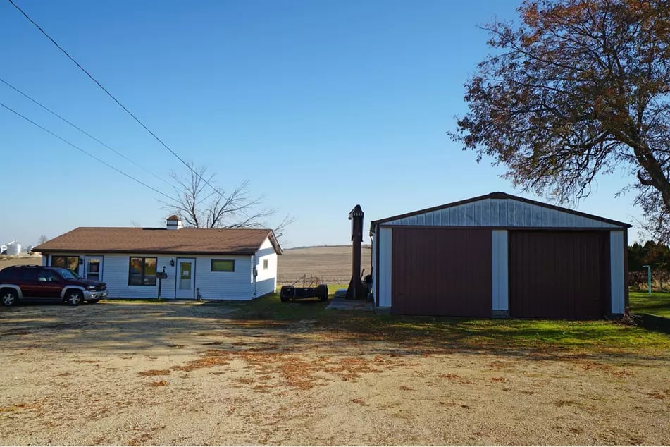 Small white house beside a large detached red-brown garage, with a gravel driveway and open fields.