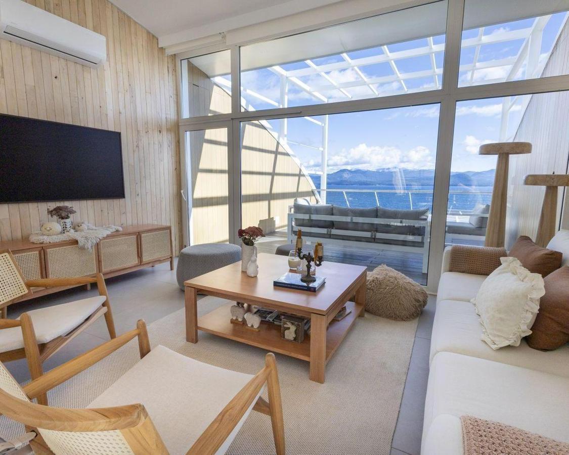 Living room with pale wood paneling and large windows overlooking water and mountains.