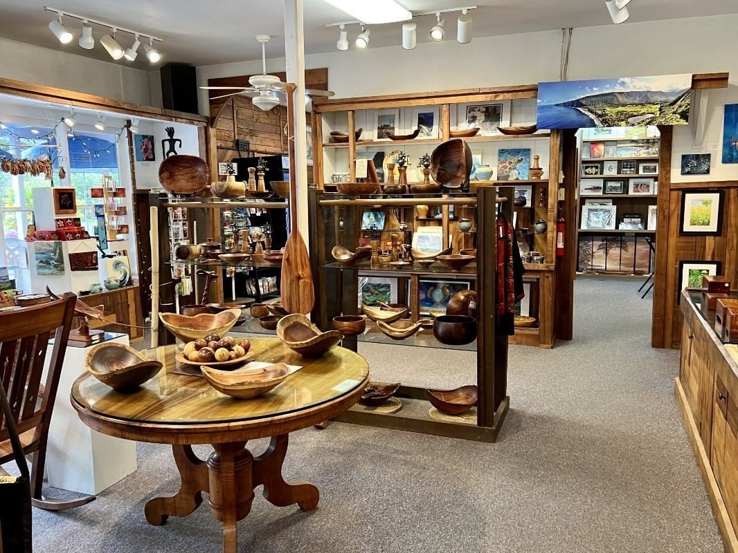 Retail shop interior with wooden shelves and tables displaying carved bowls, crafts, and framed artwork.