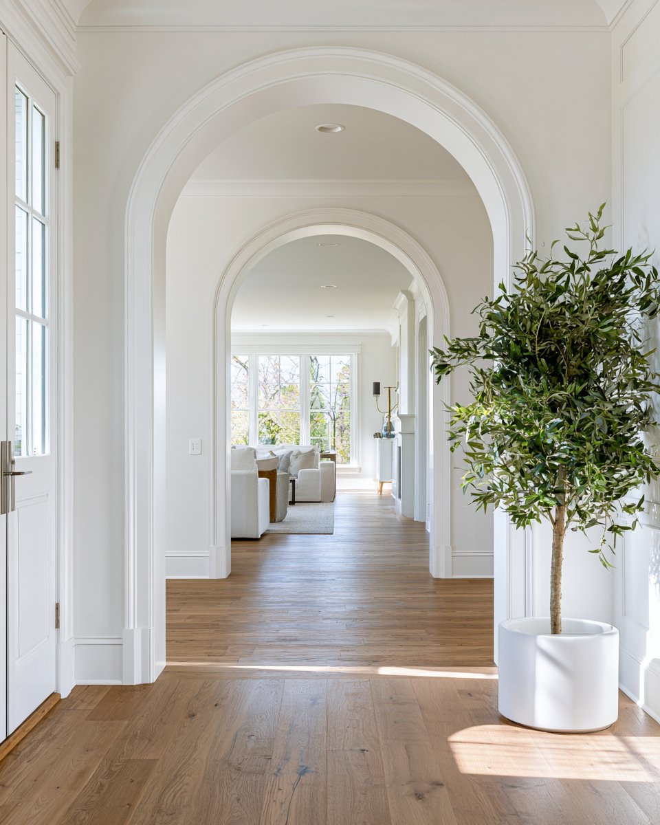 Hardwood hallway with white arched doorways, a potted tree, and a sunlit living room beyond.