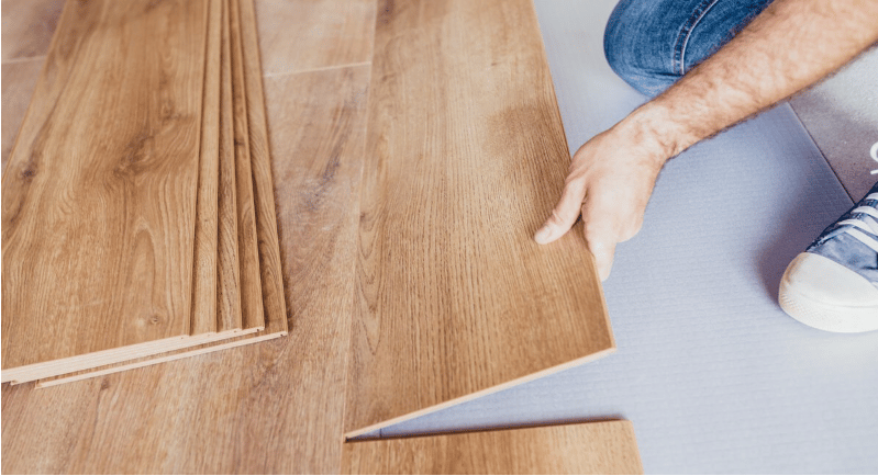 A person holds a wood-look flooring plank beside stacked flooring panels.
