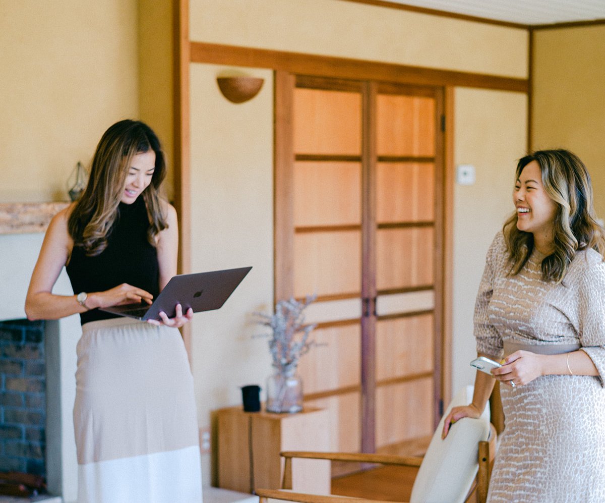 Two women smiling together in an office, one holding an open laptop.