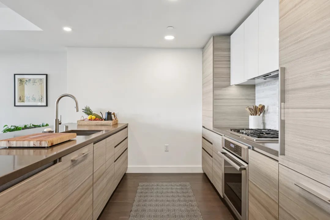 A modern kitchen with light wood grain cabinets, stainless steel countertops, and a gray patterned runner rug.