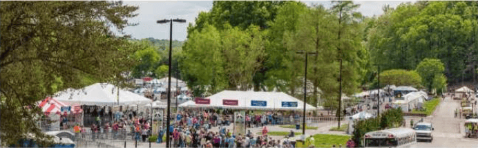 Crowds gather among vendor tents at a tree-lined outdoor fair.