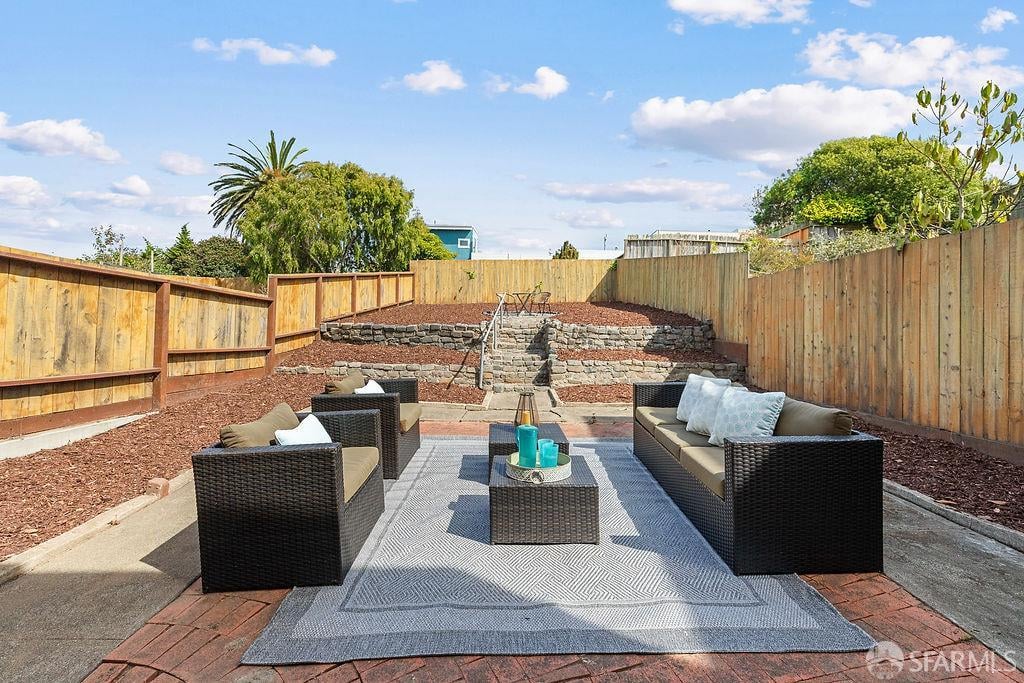 Backyard patio with wicker seating on a rug, enclosed by wooden fences and bordered by stone retaining steps.