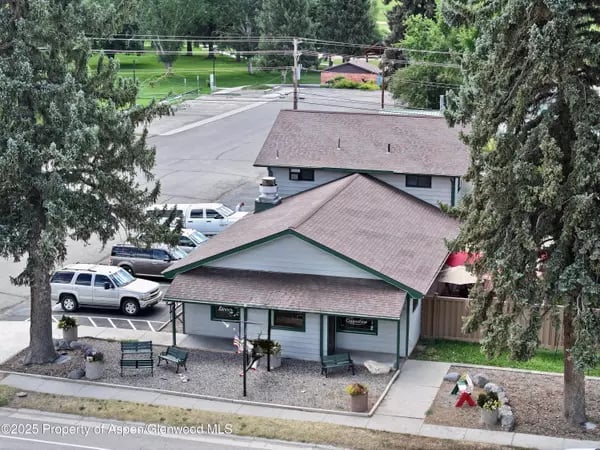 Small office building with green trim, a covered porch, parked cars, and mature trees.