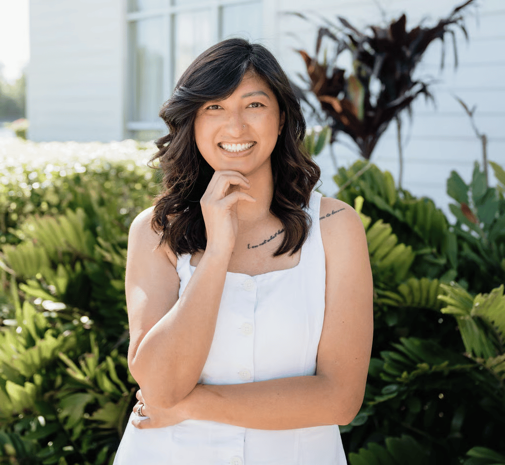 Smiling woman in a white sleeveless top, posing in front of leafy plants.
